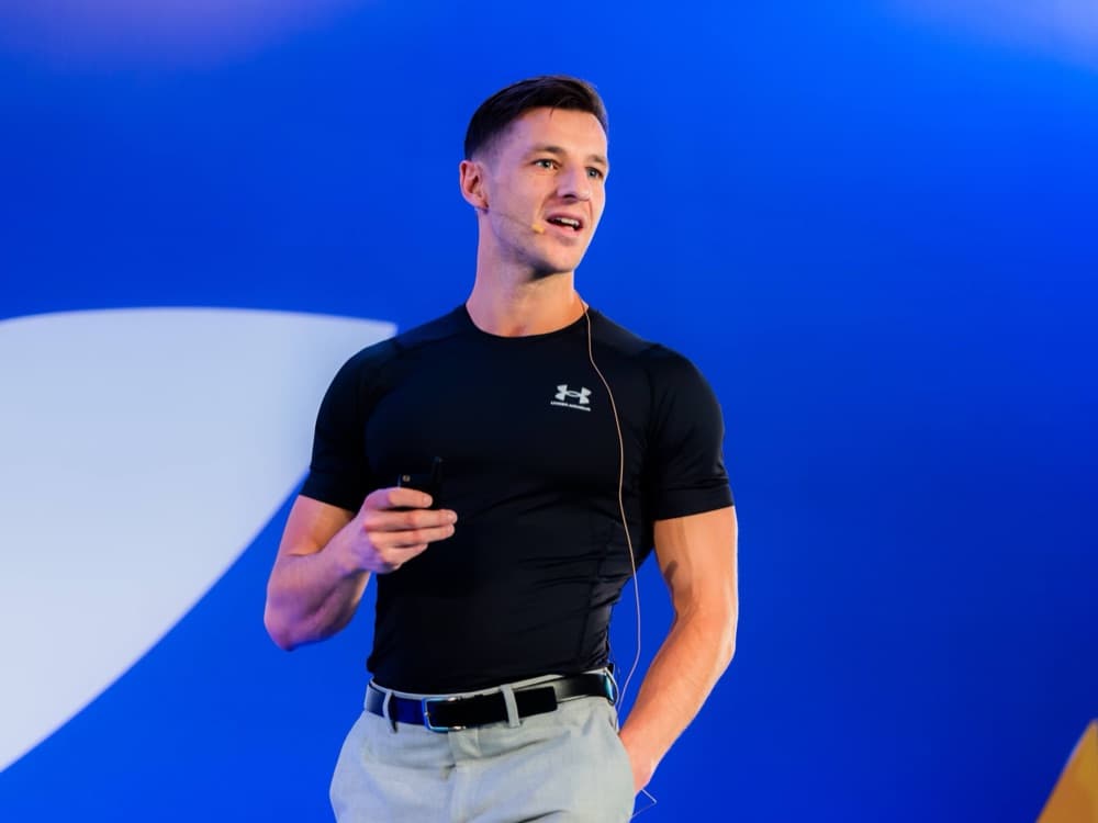 Delano presenting on a blue-lit stage, headset mic and clicker in hand